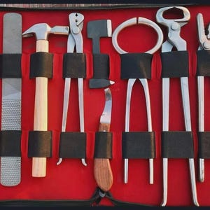Peut inclure: Une trousse à outils rouge contenant divers outils de maréchal-ferrant en métal, dont une râpe, un marteau, des pinces et un couteau à sabot. Les outils sont maintenus en place par des sangles noires. Pour le soin des sabots.