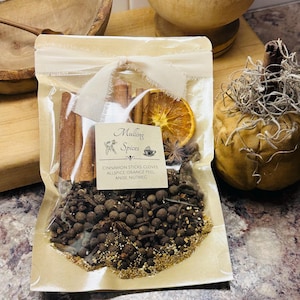 May include: A clear cellophane bag filled with mulling spices, including cinnamon sticks, cloves, allspice, orange peel, anise, and nutmeg. The bag is tied with a cream-colored ribbon and features a label with the text "Mulling Spices."