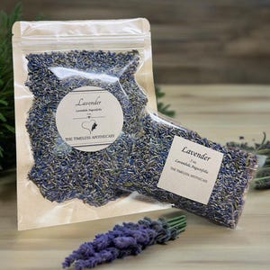 Può includere: Due sacchetti trasparenti pieni di boccioli di lavanda essiccati. Ogni sacchetto ha un'etichetta con la scritta "Lavender" e "The Timeless Apothecary". Un mazzo di fiori di lavanda fresca è in primo piano.