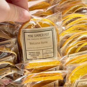 May include: A collection of individually packaged dried orange slices and cinnamon sticks labeled Mini Simmer Pot The Timeless Apothecary Welcome Home. Each clear plastic bag contains dried fruit and spices with a net weight of .5 oz.