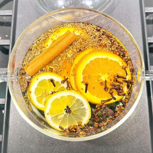 May include: A clear glass bowl filled with water, orange and lemon slices, cloves, and a cinnamon stick. The bowl is on a dark surface, and the ingredients are arranged for a fragrant infusion.
