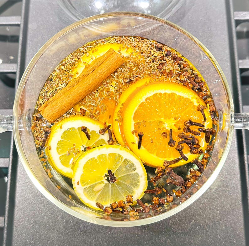 May include: A clear glass bowl filled with water, orange and lemon slices, cloves, and a cinnamon stick. The bowl is on a dark surface, and the ingredients are arranged for a fragrant infusion.