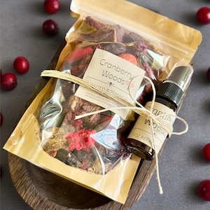 Może przedstawiać: Drewniana miska zawiera przezroczystą torbę z suszonymi roślinami i małą szklaną butelkę z rozpylaczem, obie z etykietą "Cranberry Woods". Torba i butelka są związane sznurkiem. Świeże, czerwone żurawiny są rozrzucone.