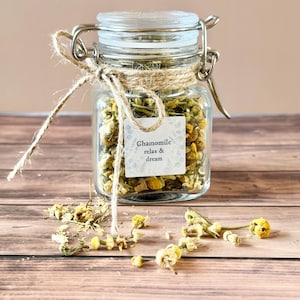 May include: A clear glass jar filled with dried chamomile flowers, secured with a metal clasp and twine. A small label reads "Chamomile relax & dream." The jar sits on a wooden surface, with loose chamomile blossoms scattered around.