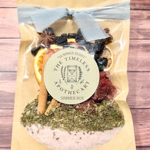 Könnte beinhalten: Ein klarer Zellophanbeutel, gebunden mit einem hellblauen Band, gefüllt mit getrockneten Kräutern, Gewürzen und Früchten. Der Beutel hat ein rundes Etikett mit der Aufschrift "Summer Berry The Timeless Apothecary Simmer Pot."