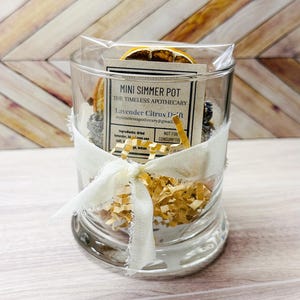 以下が含まれることがあります： 白いリボンで結ばれた、乾燥ラベンダー、柑橘類、その他の材料が入った透明なガラス瓶。瓶には「Mini Simmer Pot」と「Lavender Citrus Dream」と書かれたラベルが付いています。背景は明るい木目模様です。
