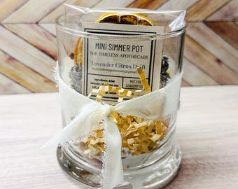 May include: A clear glass jar filled with dried lavender, citrus, and other ingredients, tied with a white ribbon. The jar contains a label that reads "Mini Simmer Pot" and "Lavender Citrus Dream". The background is a light wood pattern.