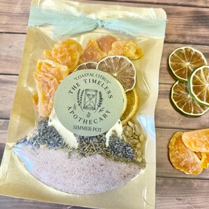May include: A clear pouch with a gold backing contains dried orange slices, lime slices, lavender, and salt. A light green ribbon ties the top. A circular label reads THE COASTAL CITRUS TIMELESS APOTHECARY SIMMER POT with an hourglass icon.