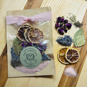 May include: A clear cellophane bag filled with dried botanicals, tied with a pink ribbon. The bag contains dried lemon slices, rose buds, lavender, and bay leaves. A green and white sticker reads "Spring Awakening The Timeless Apothecary Simmer Pot."