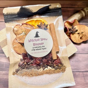 Peut inclure: Un sachet transparent contenant des tranches d'orange et de pomme séchées, des bâtons de cannelle, de l'anis étoilé, des canneberges et des épices. Une étiquette ronde indique Witches Brew Simmer, A cozy brew for crisp autumn nights, THE TIMELESS APOTHECARY.