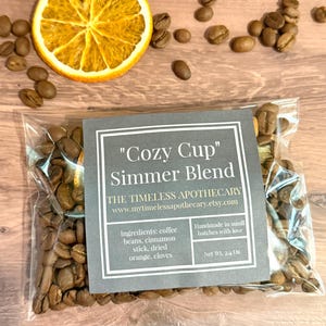 May include: A clear bag of "Cozy Cup" Simmer Blend with coffee beans. The label includes ingredients: coffee beans, cinnamon stick, dried orange, and cloves. A dried orange slice and loose coffee beans are scattered around the bag. Net weight is 2.4 oz.