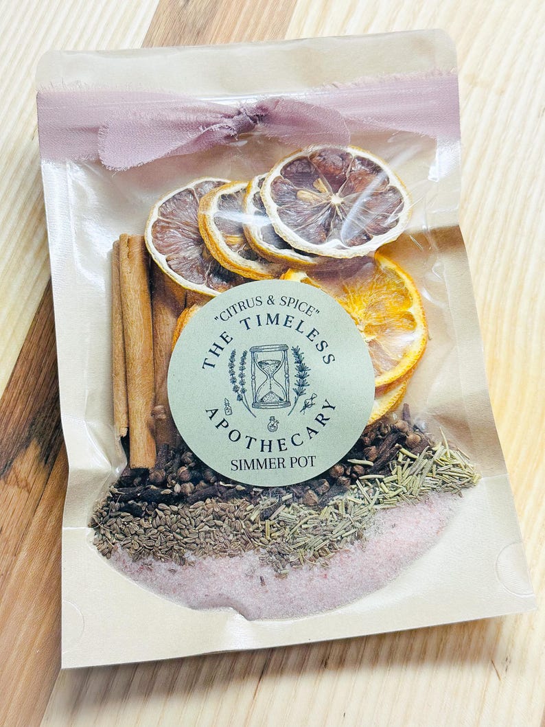 May include: A clear cellophane bag filled with dried citrus slices, cinnamon sticks, and various spices. The bag is sealed with a pink ribbon and features a round label that reads "Citrus & Spice" and "The Timeless Apothecary Simmer Pot."