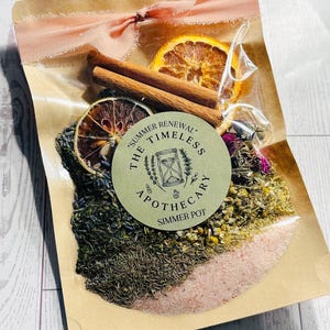 Könnte beinhalten: Ein Cellophanbeutel mit getrockneten Kräutern, Gewürzen und Zitrusfruchtscheiben, gebunden mit einem rosa Band. Der Beutel enthält Zimtstangen, getrocknete Orangenscheiben und eine Vielzahl von getrockneten Kräutern und rosa Salz. Ein runder Aufkleber lautet "Summer Renewal The Timeless Apothecary Simmer Pot."