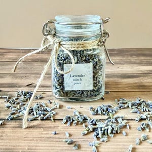 May include: A clear glass jar filled with dried lavender buds, secured with a metal clasp and twine. A label on the jar reads "Lavender calm & peace." Scattered lavender buds are on a wooden surface.
