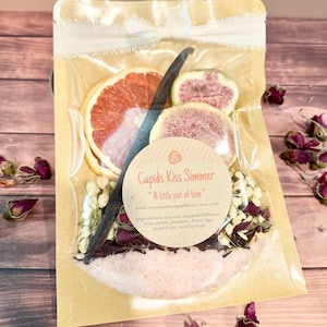 May include: A clear cellophane package tied with a white ribbon contains a bath simmer. The simmer includes dried grapefruit slices, a vanilla bean, and rose petals. The label reads "Cupids Kiss Simmer" and "A little pot of love."