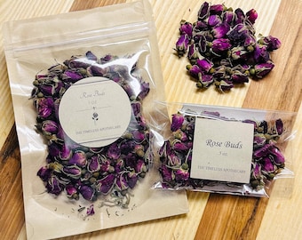Puede incluir: Dos bolsas de plástico transparente y un montón de capullos de rosa secos sobre una superficie de madera. Una bolsa contiene 28g de capullos de rosa, la otra 14g. Ambas bolsas tienen una etiqueta circular que dice "Rose Buds" y "The Timeless Apothecary". Los capullos de rosa son de color morado oscuro.