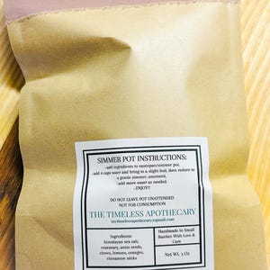 May include: A brown paper bag with a label that reads "Simmer Pot Instructions." The label lists ingredients like Himalayan sea salt, rosemary, and cinnamon sticks. The bag is sealed with a pink ribbon. Net weight is 3 oz.
