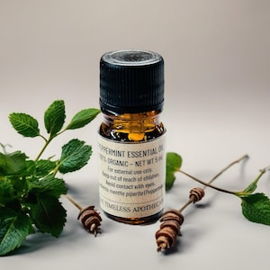 Può includere: Una piccola bottiglia di vetro ambrato di olio essenziale di menta piperita con un tappo nero. L'etichetta recita "Peppermint Essential Oil 100% Organic - NET WT 5 ml." Foglie di menta fresca e baccelli secchi sono disposti intorno alla bottiglia.