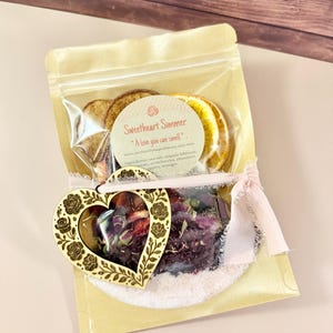 May include: A beige package containing dried orange slices, a heart-shaped wooden ornament, and a sachet of dried flowers. The ornament is decorated with rose designs. The package is tied with a pink ribbon and includes the text "Sweetheart Simmer".