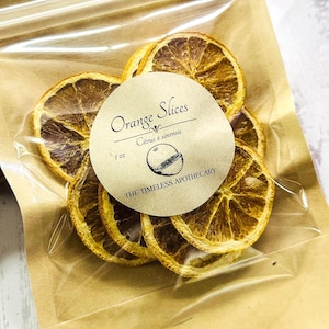 May include: A clear plastic bag filled with dried orange slices. A round label reads "Orange Slices Citrus x sinensis" and "THE TIMELESS APOTHECARY". The bag is sealed with a brown paper backing. The orange slices are a warm brown color.