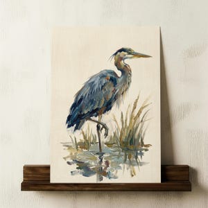 May include: A canvas print depicting a blue heron in water, with reeds. The bird is painted in shades of blue and grey, with a long beak and detailed feathers. The artwork is displayed on a dark wooden shelf, adding a touch of nature.