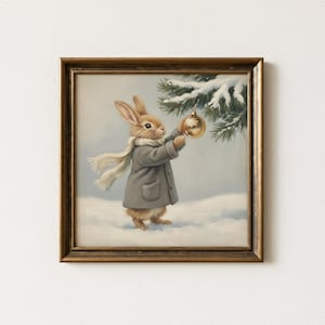 Puede incluir: Una impresión enmarcada representa un conejo con abrigo y bufanda grises, decorando un árbol de Navidad nevado con un adorno dorado. La obra de arte tiene una estética vintage, con un marco dorado y una paleta de colores invernal suave.