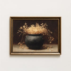 Op de afbeelding: Een ingelijst stilleven schilderij met een zwarte keramische pot gevuld met gedroogde, beige bloemen. Het schilderij heeft een donkerbruine achtergrond en een goudkleurige lijst. Het kunstwerk heeft een vintage, rustieke uitstraling.