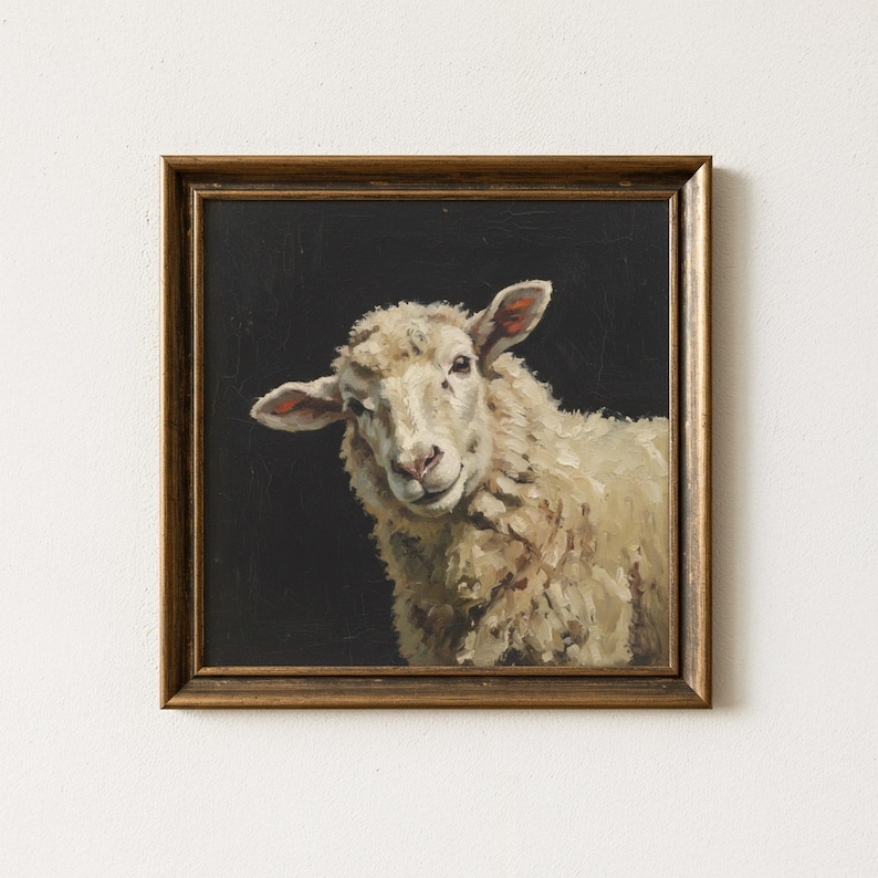 Op de afbeelding: Een ingelijst schilderij van een schaap met een lichtgekleurde wollen vacht en roze neus en oren. Het schaap staat tegen een zwarte achtergrond. De lijst heeft een donkerbruine, warme kleur.
