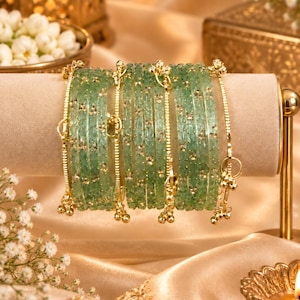 May include: A set of translucent light green glass bangles featuring embedded gold-tone floral accents and small dangling gold-tone charms. The circular bracelets are arranged in a row, showcasing a textured surface with metallic gold detailing.