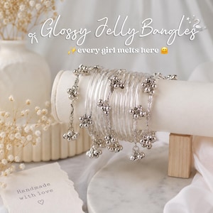 May include: Transparent jelly bangles with silver floral charms. The image features the text "Glossy Jelly Bangles" and "every girl melts here." A card with the words "Handmade with love" is also visible.
