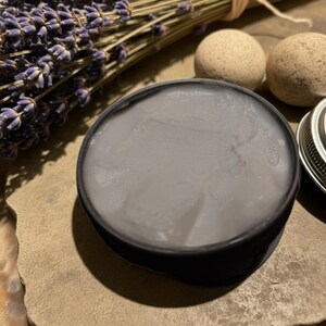 Puede incluir: Un tarro negro lleno de crema gris claro, con un manojo de lavanda seca y dos peque&ntilde;as piedras beige redondas en el fondo. La tapa del tarro es parcialmente visible a la derecha. La crema tiene un aspecto liso y ligeramente texturizado.