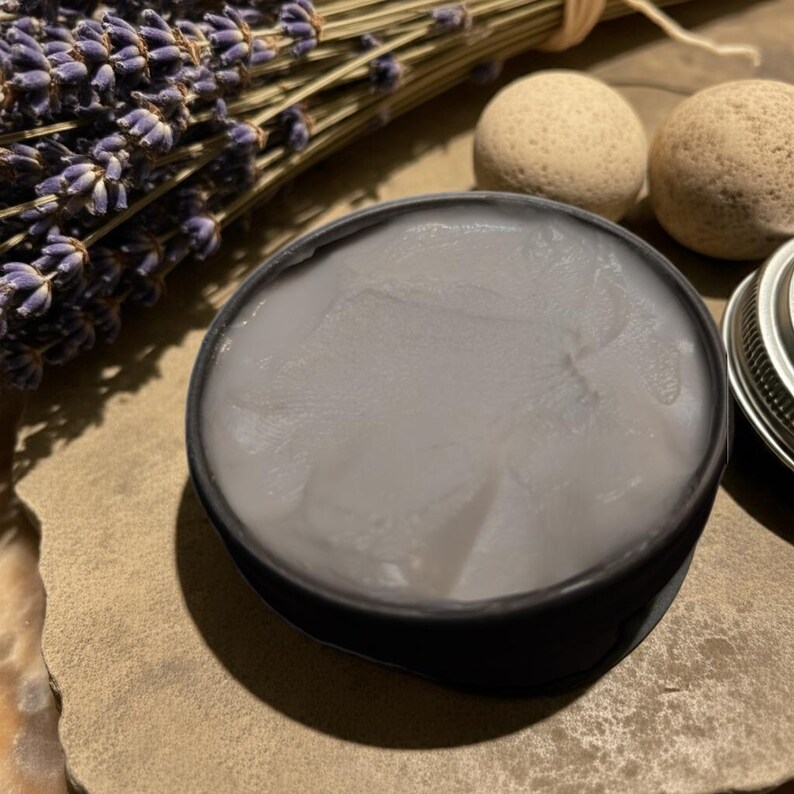 Puede incluir: Un tarro negro lleno de crema gris claro, con un manojo de lavanda seca y dos peque&ntilde;as piedras beige redondas en el fondo. La tapa del tarro es parcialmente visible a la derecha. La crema tiene un aspecto liso y ligeramente texturizado.