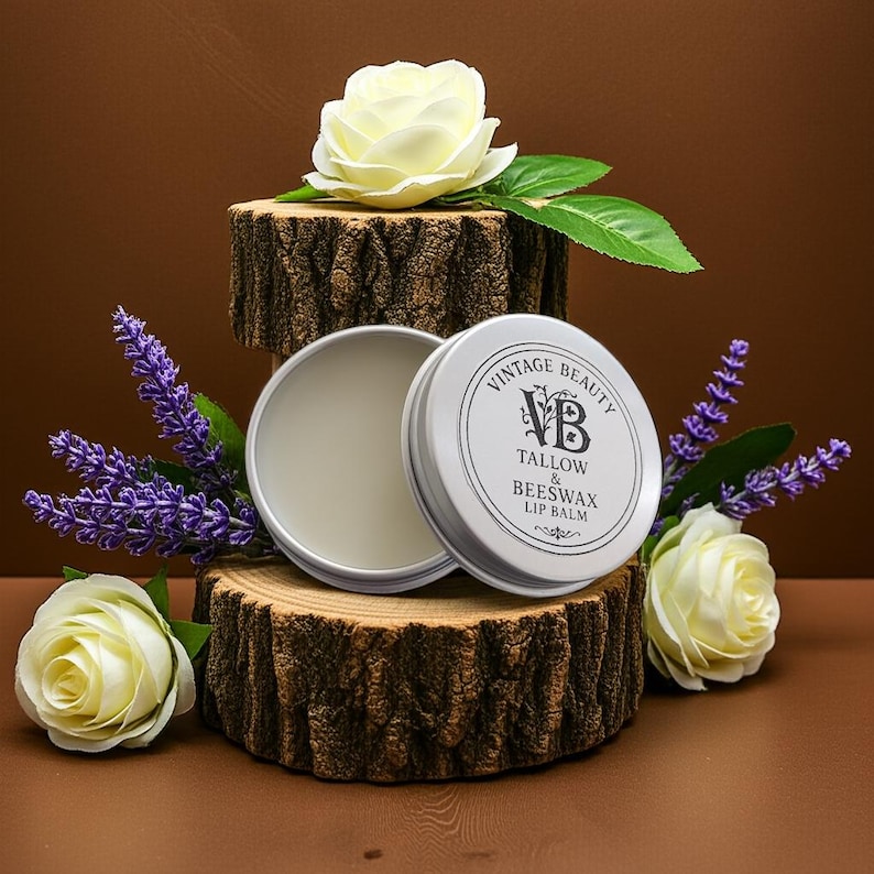 Puede incluir: Un bote plateado de b&aacute;lsamo labial Vintage Beauty Tallow & Beeswax, abierto. El bote est&aacute; sobre una rodaja de madera, rodeado de rosas blancas y lavanda morada. El fondo es un degradado marr&oacute;n.
