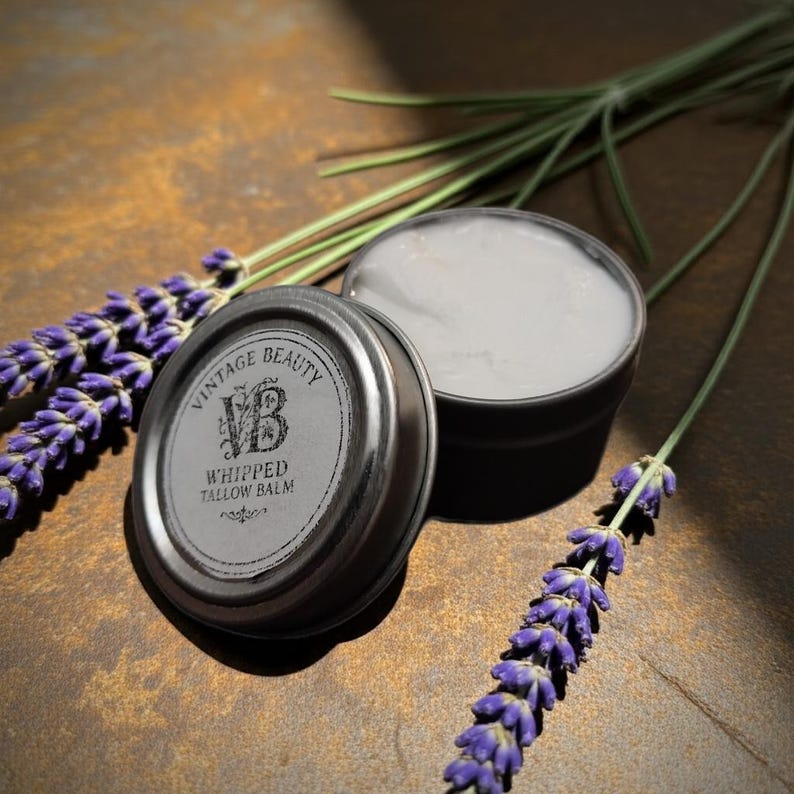 Puede incluir: Una lata de Vintage Beauty Whipped Tallow Balm con la tapa quitada, revelando el b&aacute;lsamo. La tapa est&aacute; al lado de la lata. El texto en la tapa dice "Vintage Beauty Whipped Tallow Balm". Ramas de lavanda est&aacute;n a un lado.
