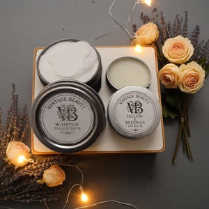 Puede incluir: Una colecci&oacute;n de productos para el cuidado de la piel Vintage Beauty, que incluyen b&aacute;lsamo de sebo batido y b&aacute;lsamo labial de cera de abejas de sebo, en peque&ntilde;as latas de metal. Las latas est&aacute;n dispuestas sobre una bandeja blanca, con flores secas y luces de hadas en el fondo.