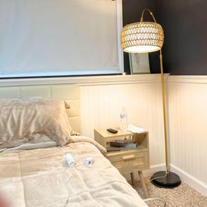 May include: A woven rattan floor lamp with a gold-colored metal pole and a black circular base. The lamp has a cylindrical shade and is positioned next to a light-colored wooden nightstand and a bed with a beige comforter.
