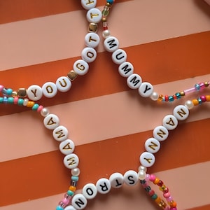 May include: Three circular beaded bracelets featuring white round beads with gold lettering. The bracelets display the phrases YOU GOT THIS, MUMMY, MAMA, NANNY, and STRONG. The designs incorporate small colourful glass beads in shades of pink, blue, orange, and gold.