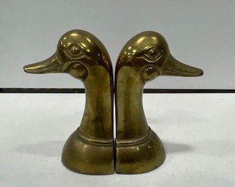 May include: A pair of polished brass bookends shaped like duck heads. Each piece features a long, slender neck and a detailed duck face with defined eyes and a beak. The brass has a warm, metallic gold tone with a smooth, reflective surface finish.
