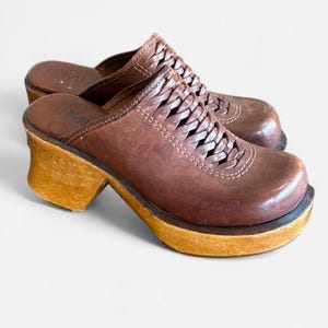 May include: A pair of brown leather mules featuring a woven strap design across the top, a rounded toe, and a thick wooden platform heel. The interior footbed is brown leather with the brand name MIA printed in a dark font on the heel area.