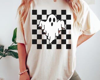 May include: A cream-coloured t-shirt with a white ghost graphic set against a black and white chequered pattern. The ghost has a simple design with two eyes and an open mouth. The shirt has short sleeves and a relaxed fit.