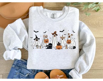 May include: A light gray sweatshirt featuring a Halloween-themed design. The graphic depicts several dogs dressed as ghosts, witches, and holding jack-o'-lanterns. Black bats are scattered across the top. The sweatshirt is displayed on a denim surface.