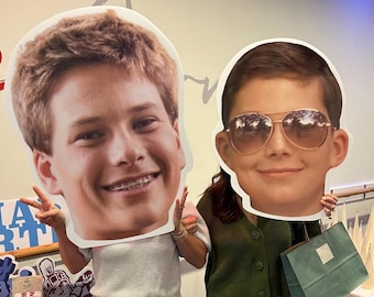 May include: Two large cardboard cutouts of people's faces. The person on the left is smiling and wearing braces. The person on the right is wearing sunglasses and smiling. A person is holding each cutout.