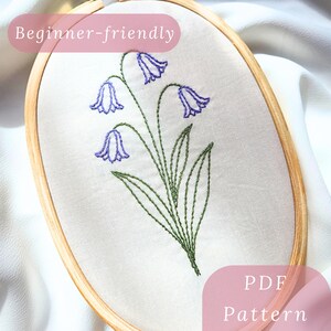 以下が含まれることがあります： 刺繍枠に花の模様。白い布に緑色の茎と葉を持つブルーベルの花が刺繍されています。「Beginner-friendly」と「PDF Pattern」の文字が見えます。