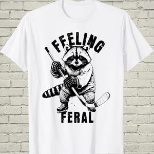 May include: White t-shirt featuring a black and white illustration of a raccoon playing hockey. The raccoon is wearing a jersey and holding a hockey stick. The text "I FEELING" is above the raccoon, and "FERAL" is below.