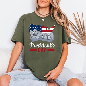 May include: Olive green t-shirt featuring a graphic of Mount Rushmore within a map of the United States, with the American flag incorporated. The text "President's Day" is displayed below the graphic in white and red.