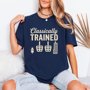 May include: Navy blue t-shirt with the text "Classically Trained" in a cream-colored font. Below the text are illustrations of a gear shift, pedals, and a stick shift. The shirt is a casual style.