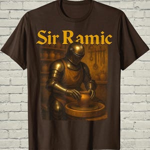 May include: A dark brown t-shirt featuring a graphic of a knight in full metal armor wearing an apron while working at a pottery wheel. The text Sir Ramic is printed in yellow above the knight. The scene includes shelves with pottery.