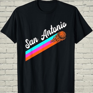 May include: Black t-shirt featuring the text "San Antonio" in white script, with a basketball graphic and colorful diagonal stripes. The basketball has flames trailing behind it.