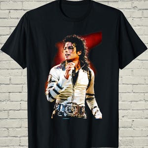 May include: Black t-shirt featuring a vibrant image of Michael Jackson performing on stage. The graphic shows him wearing a white jacket, a decorative belt, and holding a microphone. The background is a red abstract shape.