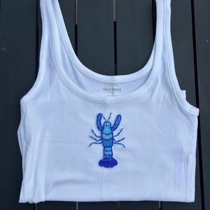 May include: White ribbed tank top with a blue beaded lobster design. The tank top has a scoop neck and is made of a soft, stretchy material. The lobster design is centered on the front of the tank top.